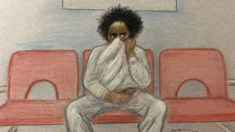 Julia Quenzler Court sketch showing Axel Rudakubana sat in the middle of a row of three red chairs. He's wearing a grey tracksuit, holding an item in his left hand and his right hand is holding his sweater onto his face - hiding his nose and mouth