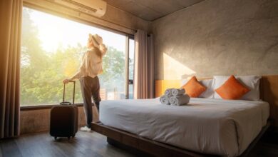 A woman standing next to her suitcase and looking out the window of her sunny hotel room.