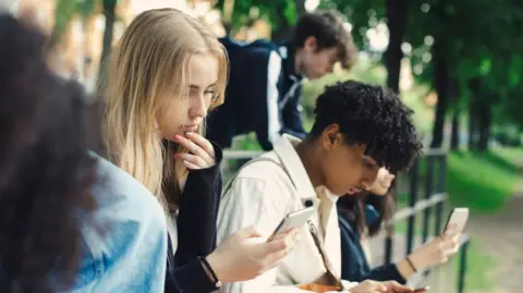 Getty Images A group of teens look down at their smartphones in a park.