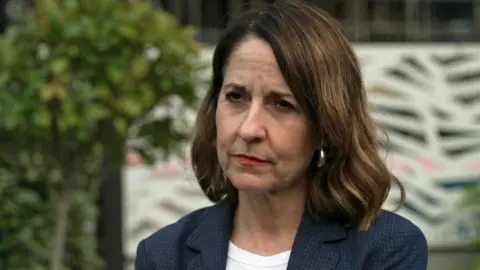 BBC A medium close up of Pensions Secretary Liz Kendall wearing a blue jacket and white top with tree and bushes in background