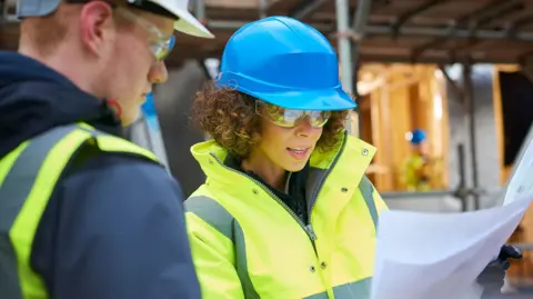 Getty Images A man and woman wearing hardhats and hi-vis clothing looking at building plans on a building site