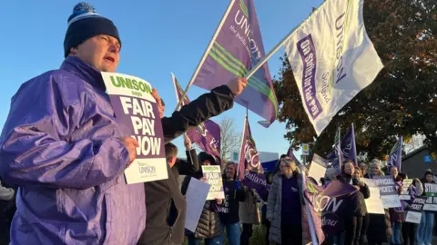 BBC Man in purple jacket and bobble hat holds fair pay now sign as a crowd of picketers behind him wave flagsand banners under a clear blue morning sky.