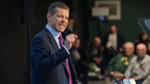 Plaid Cymru Rhun ap Iorwerth speaking at his party conference, in a dark suit, a blue shirt and a purple pokadot tie, in front of a teleprompter.