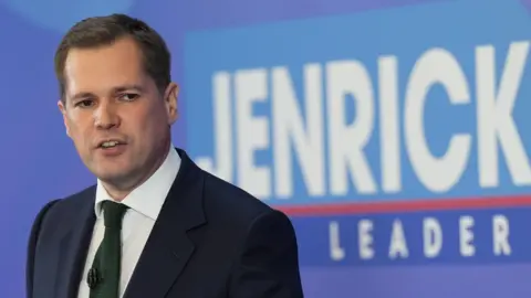 PA Media Tory leadership hopeful Robert Jenrick speaks to supporters as he stands in front of a sign saying 