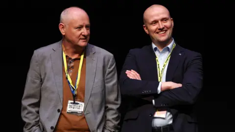 Getty Images Murray Foote and Stephen Flynn - Mr Foote is wearing a grey jacket and brown shirt with a lanyard around it, while Flynn has his arms folded and is smiling