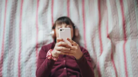 Getty Images Girl in bed with earphones on looking at her phone
