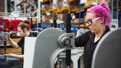 Getty Images A woman with pink hair uses a hole punch machine in a factory making bicycles, with another worker in the background in a manufacturing environment.