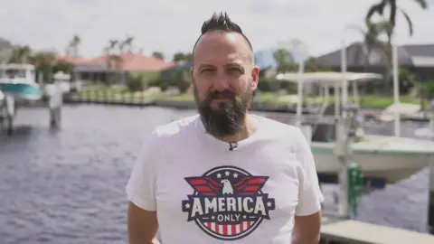 Free, a man with a full beard and closely cropped brown hair with a quiff on top, wearing a T-shirt with a stylised eagle and the slogan 