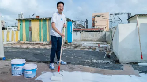 i2Cool Martin Zhu painting the flat roof of a building with a long-handled roller and his firm's white paint.