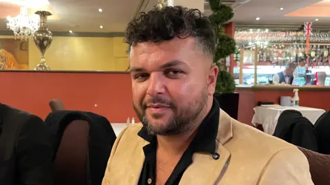 Ben Schofield/BBC Alex Rafiq, looking direct to camera, seated in a restaurant. He has curly, black hair, cut shorter at the sides and longer on top, and dark eyes. He is wearing a camel coloured jacket and black shirt, open at the collar. Over his shoulder on the right of the image is a bar area where a member of staff can be seen working. 