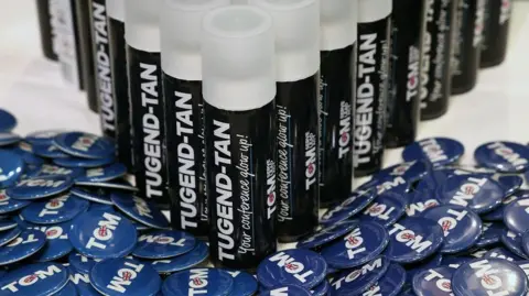 Getty Images Cans of promotional Tugend-tan lined up on a bed of Tom Tugendhat badges at the Tory conference in Birmingham