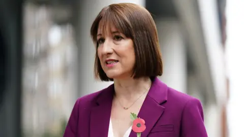 PA Media Rachel Reeves, with dark hair and wearing a purple suit, speaks with a blurred out concrete structure behind her 