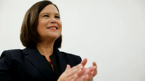 Reuters Mary Lou McDonald - a woman with short brown hair above her shoulders applauds while standing against a white background. She is wearing a dark suit jacket and a red, beaded necklace.