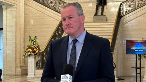 Conor Murphy standing at a microphone in the great hall at Stormont wearing a suit and tie. Behind him a grand staircase. 