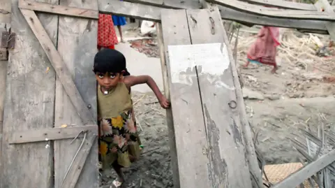 Getty Images A young Bangladeshi girl wearing a green dress with flowers on the skirt stands in the wooden doorway of her collapsed house. 