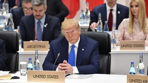 Getty Images Donald Trump attends the G20 Summit in Osaka, Japan in 2019. He sits behind a United States sign, with his fingertips touching, and wears a blue suit with a blue tie. His daughter Ivanka Trump is visible at a desk behind his right shoulder. She has long blonde hair and wears a pale pink long-sleeved dress. 