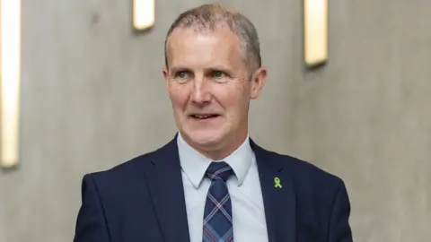 PA Media Michael Matheson, with thinning grey hair and wearing a dark blue suit, light blue shirt and blue tartan tie, looks to his right while walking in the Scottish Parliament 