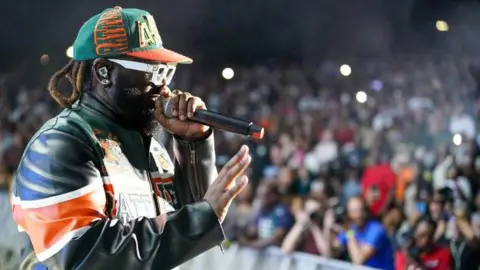 Getty Images T-Pain, pictured wearing a colourful leather jacket, white sunglasses and a green and orange baseball cap, performing in Florida last weekend