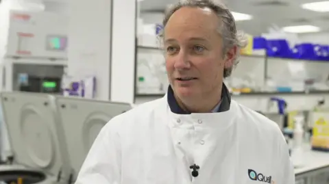 Iain McGill, chief executive of Quell Therapeutics, standing in a lab wearing a white buttoned up coat saying Quell on it