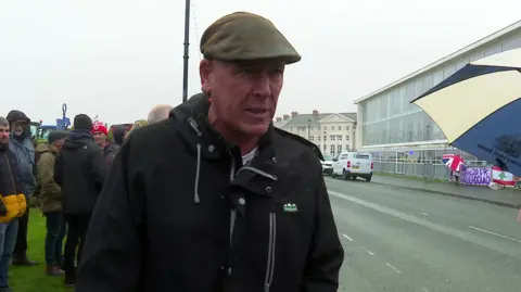 Gareth Wyn Jones standing the otherside of the road from Venue Cymru, where the Welsh Labour conference is taking place. He is wearing a flat cap and a rain coat and there are other protesters behind him and the sky is grey