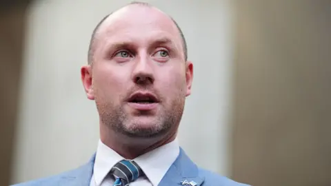 PA Media Neil Gray photographed from the shoulders up wearing a light blue suit, white shirt and blue, black and grey striped tie