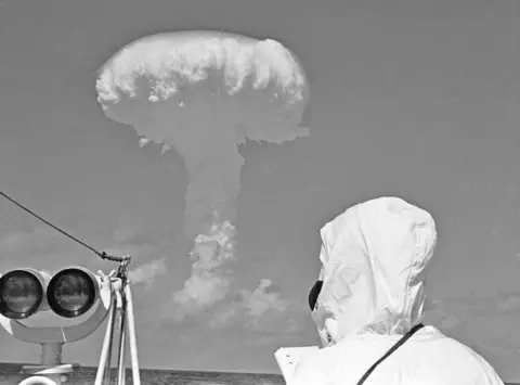 Getty Images A mushroom cloud rises over the sea while someone looks on from a ship, wearing a white hooded coat and black mask, next to some binoculars. HMS Alert was positioned 35 miles offshore of Malden Island, Kiribati, the site chosen for a series of nuclear tests on 31st May 1957. 