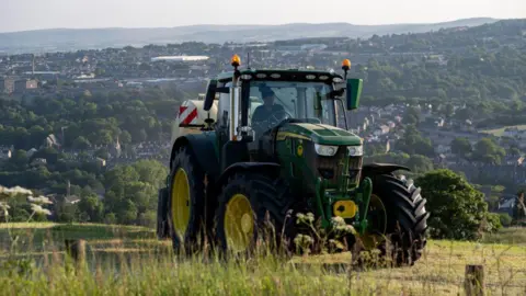 Getty Images A green tractor drives through a field, with a view of a valley in the background.