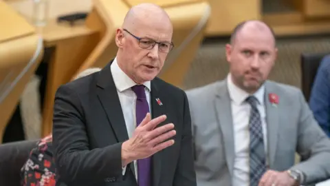 PA Media John Swinney, a bald man with glass, wearing a dark suit and purple tie, stands to speak in the Scottish Parliament 