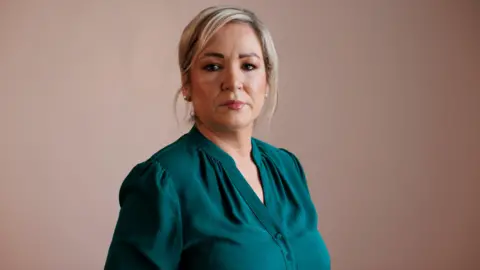 Liam McBurney/PA Wire Michelle O'Neill looking to camera with a serious expression.   She has blonde hair tied back and is wearing earrings, lipstick and a dark green blouse.