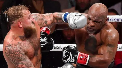 Getty Images Jake Paul, to the left of the frame, lands a punch on Mike Tyson during their so-called boxing match. The event was Netflix's first foray into live boxing. Paul is wearing sliver gloves and Tyson's gloves are black with white markings on them, similar to his infamous 'tribal' facial tattoo.