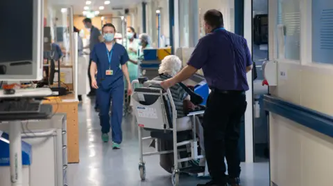 PA An NHS hospital worker, wearing a surgical mask and scrubs, walks down a busy hospital corridor 