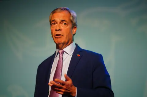 PA Media Nigel Farage wearing a blue suit, with a light blue shirt underneath and a pink tie. He is about to speak and has a mic on his face.