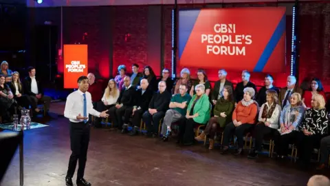 GB News Former Prime Minister Rishi Sunak, wearing a suit with a white shirt and blue tie, appears in front of an audience of people and a sign which reads 