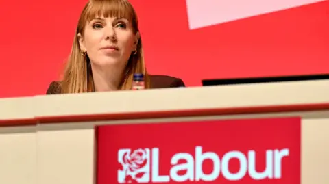 PA Media Labour Deputy Prime Minister Angela Rayner at the Labour Party Conference. She is sitting at a long table behind a red Labour logo, and in front of a red backdrop