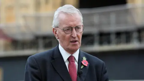 PA Media Hilary Benn has white hair and wears thin black glasses. He is wearing a black suit with a red tie. He has a red poppy on the left side of his blazer.