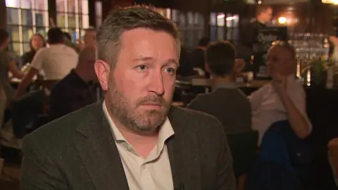 BBC Anthony Pender, owner of two pubs and a restaurant, wearing a suit while sitting in his pub