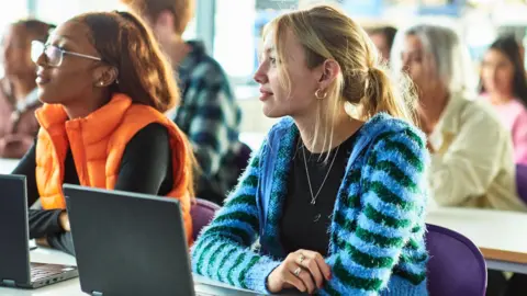 Getty Images Two female students sit at a desk with laptops open. The student in the foreground on the right is wearing a bright blue cardigan over a black T-shirt, while the student on the left has glasses and a bright orange gilet. 