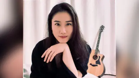 Snow Rui A woman dressed in black sits in front of a white curtain, her chin resting on the wrist of her hand. An acoustic guitar sits on a stand in the background.