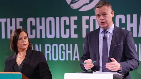 PA Media Mary Lou McDonald with Pearse Doherty who is tall with greying dark hair and is wearing a dark suit and speaking at the podium of an election event