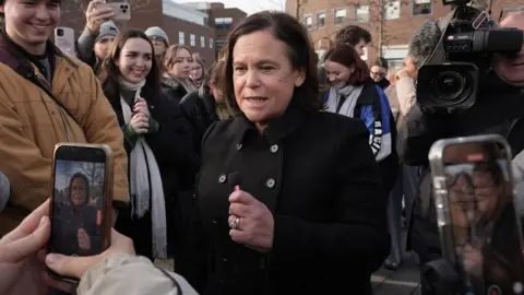 PA Media Mary Lou McDonald wearing a black button-up coat at an election event in Dublin.  Two people are taking pictures of her with their phones while a cameraman is behind her. 