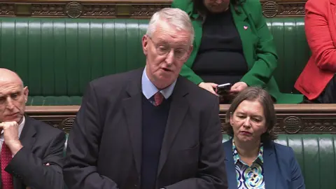 Houses of Parliament Hilary Benn speaking to the House of Commons. He is wearing a grey suit, blue shirt and red tie. He has glasses and grey hair. Behind him are other members of parliament on green leather seats.