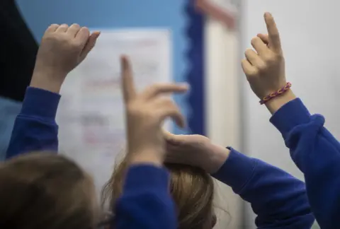 PA Young schoolchildren in blue sweatshirts raising their hands in class. One of them is wearing a loom band bracelet