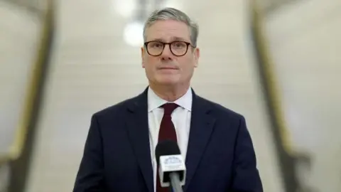 Reuters Sir Keir Starmer is wearing a navy suit with a red tie and white shirt. He is looking straight at the camera with a straight facial expression. He is wearing dark coloured glasses and has grey hair.