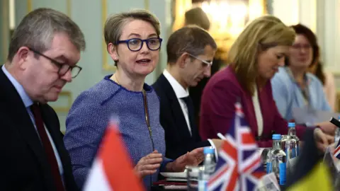 PA Media Yvette Cooper speaking around table. There are four other people seen sat on her side of the table, with small flags sitting on top.