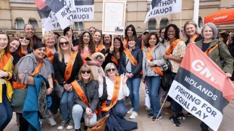 GMB Union A large group of women outside a sandstone building. All those pictured are wearing orange sashes and a woman at the front holds a large flag which says 