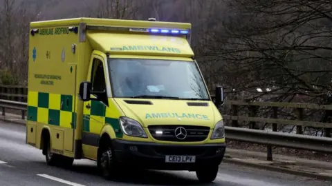 Getty Images A picture of an ambulance driving down a road. The ambulance is yellow and can be seen with the Welsh word ambiwlans written on the front.