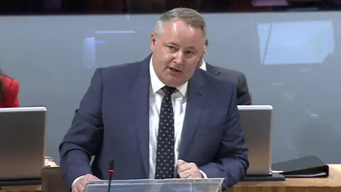 Darren Millar stood in the Senedd speaking during First Minister's Questions, wearing a blue suit and dotted tie.