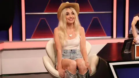 Getty Images Actress Chloe Fineman sits in a TV set on a white chair, wearing a cowboy hat, white vest top, denim shorts and silver cowboy boots. She is impersonating Hawk Tuah girl Haliey Welch. 