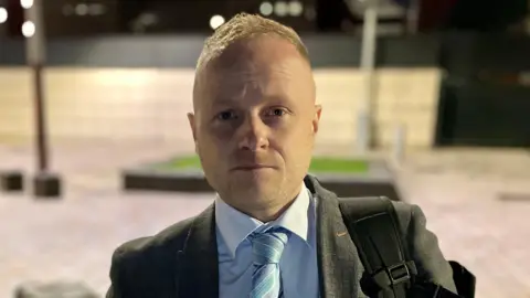 PA Media Jamie Bryson looking directly at the camera. He has short blonde hair, wearing a grey blazer, light blue shirt and blue striped tie, and carrying a backpack.