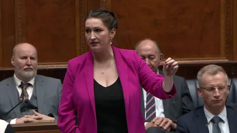 Emma Little-Pengelly speaking in the assembly. She has dark hair tied back, wearing a pink blazer, black dress, a necklace and earrings.